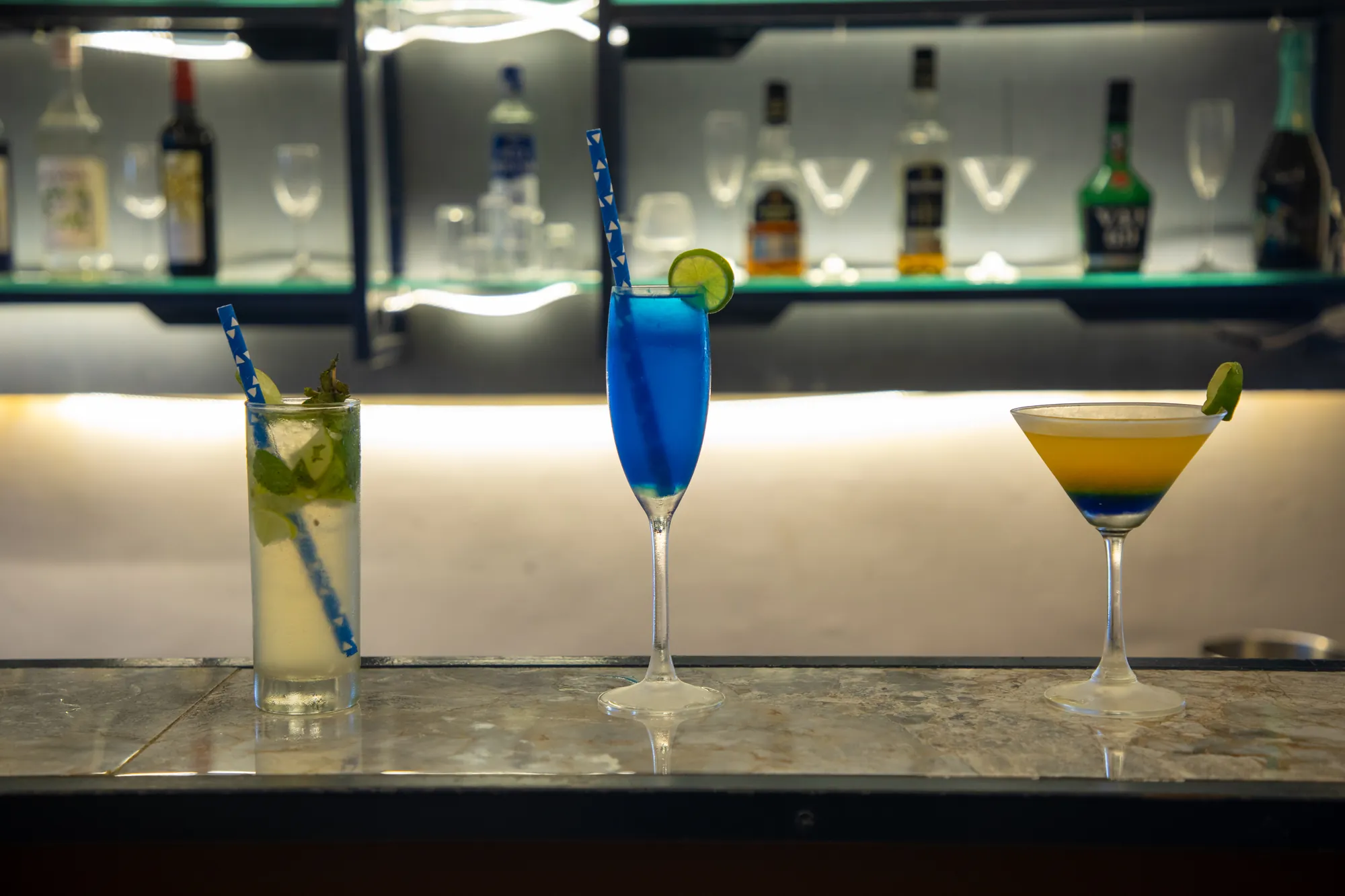 Three cocktails on the bar top: a mojito, a blue cocktail and a layered martini